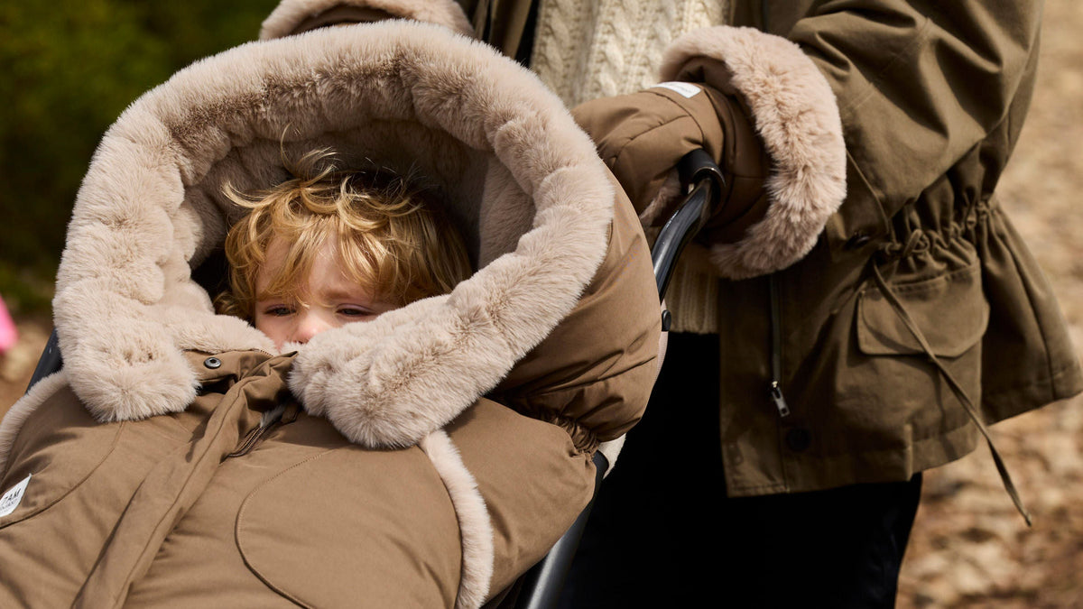 Child in a brown winter coat with fur-lined hood being pushed in a stroller by an adult.